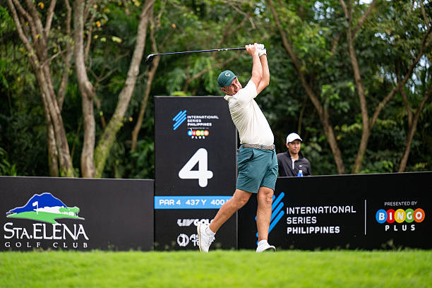 Richard Bland tees off at the 4th hole of Sta. Elena Golf Club during the International Series Philippines. He wears a light beige polo, dark green shorts, and a matching cap while finishing his powerful swing. He wears a light beige short-sleeved polo shirt. It’s breathable, moisture-wicking, and neatly designed. The athletic fit allows smooth swing movement. He pairs it with dark green golf shorts. They’re stretchable, knee-length, and comfortable for play. He wears a dark green cap with a subtle logo. It matches his shorts and provides sun protection. He sports clean white golf shoes with spiked soles. A white belt and glove complete the look.  Classic and coordinated, perfect for tournament play in the sun.
