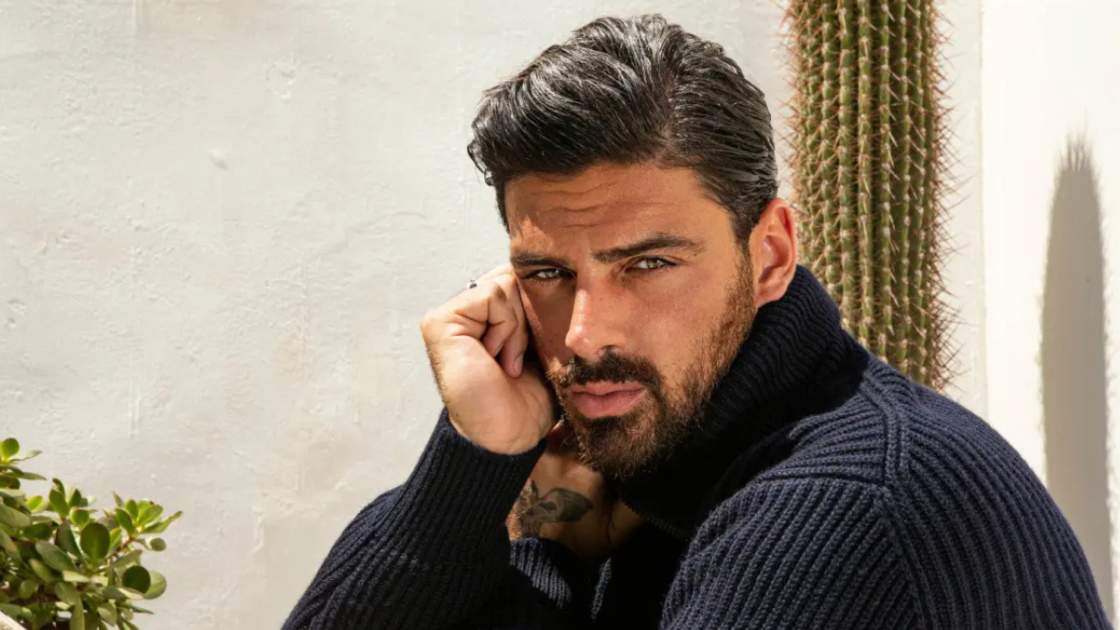 Michele Morrone posing in a navy blue ribbed turtleneck sweater for a fashion spotlight featured in 'The Housemaid,' set against a bright outdoor backdrop with desert plants.