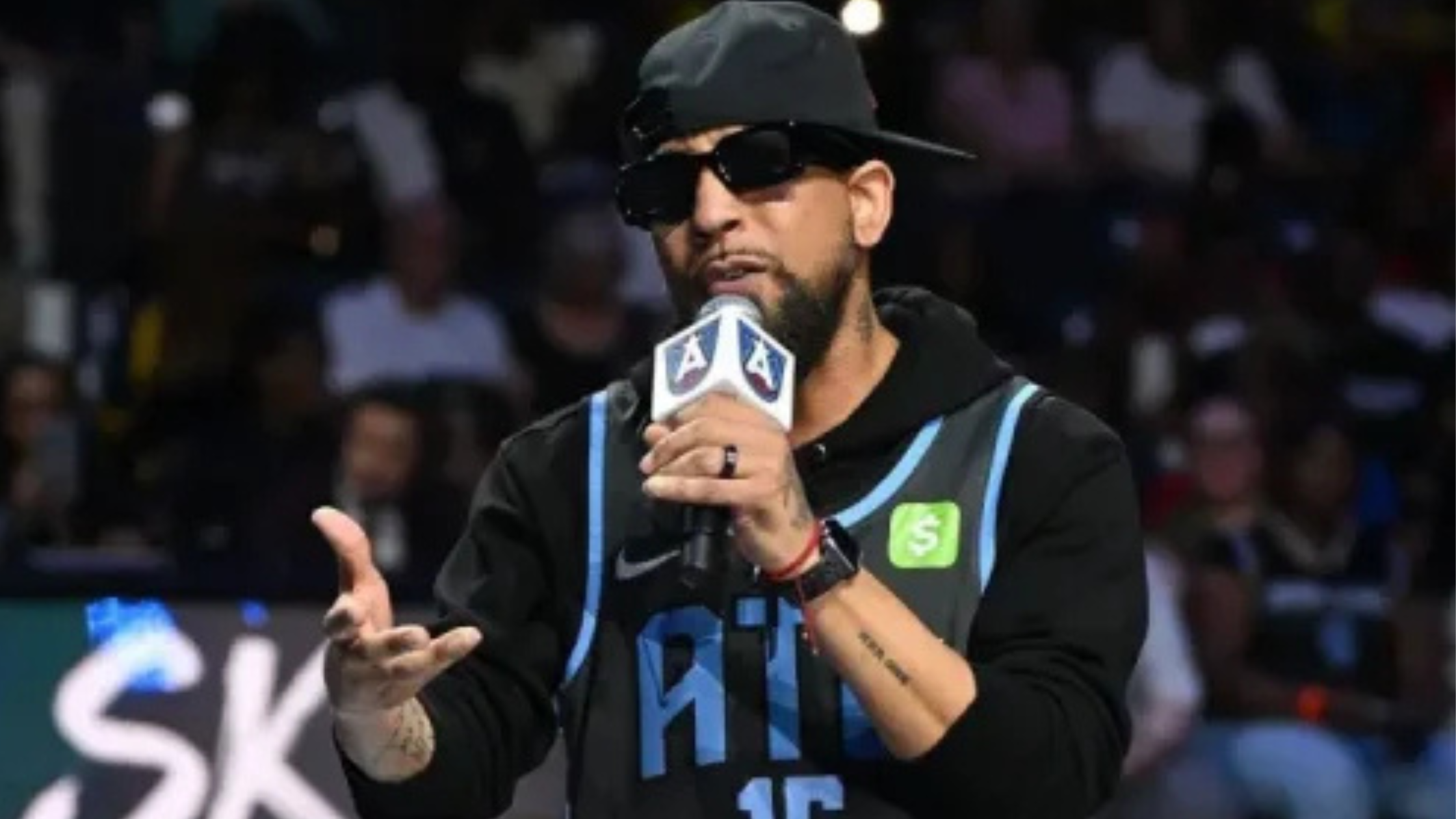 J. Holiday wearing a black ATL basketball jersey with light blue lettering, number 15, and multicolored gradient side panels during a live performance