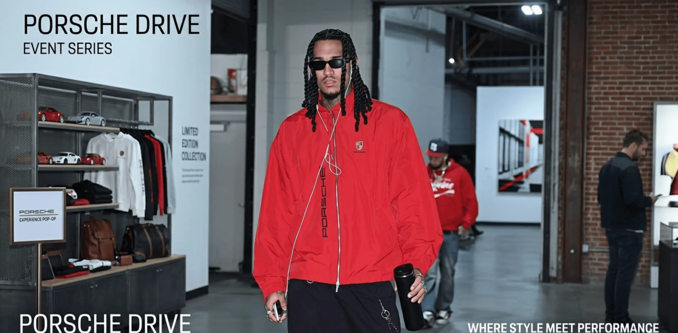 Jordan Clarkson wearing an oversized cherry-red Porsche windbreaker jacket with silver zipper and crest, styled with wired headphones at an NBA game courtside in 2026