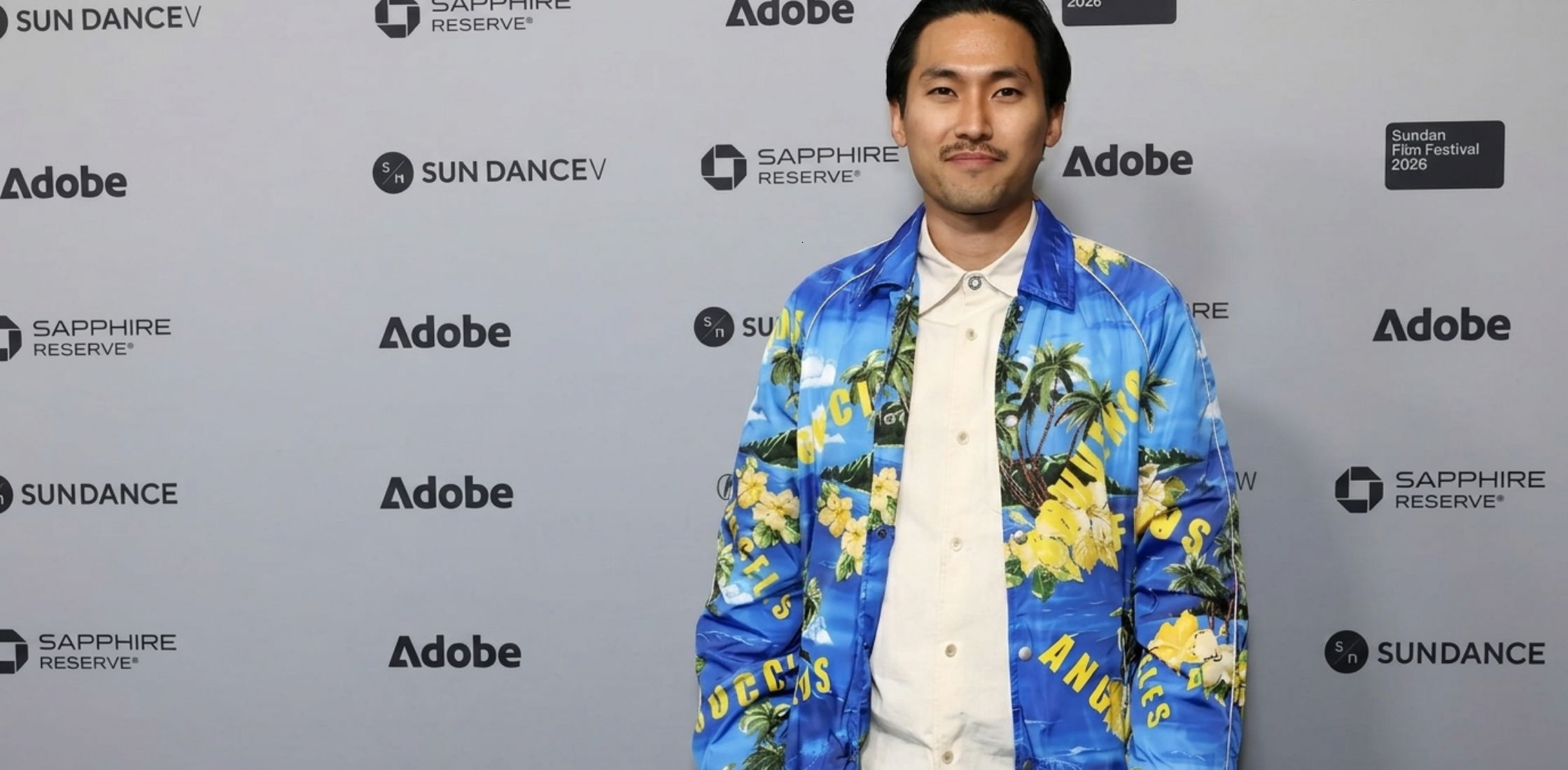 Jin Ha 2026 Sundance Film Festival blue coach jacket featuring tropical palm tree and hibiscus print, bold yellow typography, silver snap buttons, and sporty white piping with cinematic streetwear style.