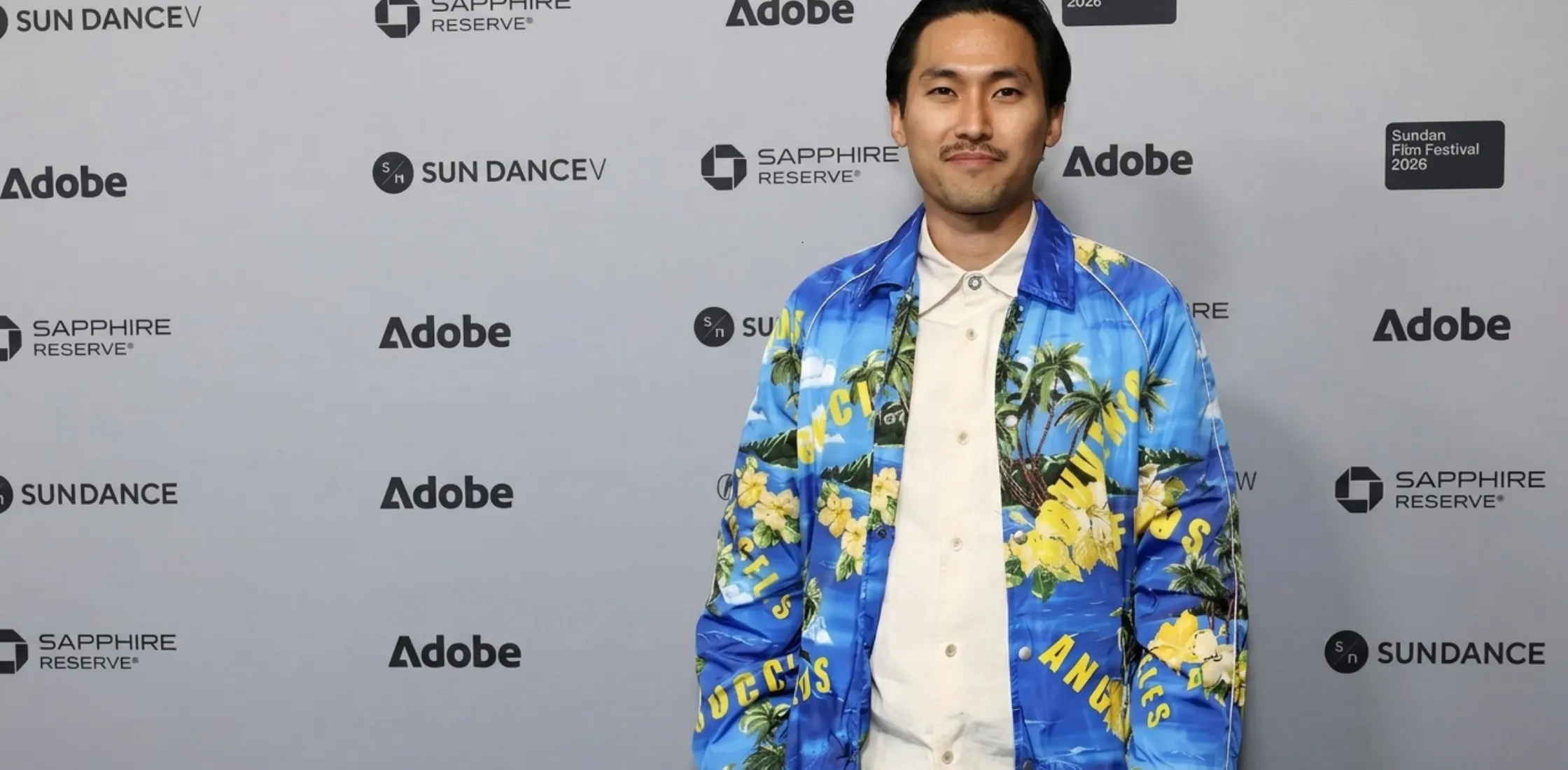 Jin Ha 2026 Sundance Film Festival blue coach jacket featuring tropical palm tree and hibiscus print, bold yellow typography, silver snap buttons, and sporty white piping with cinematic streetwear style.