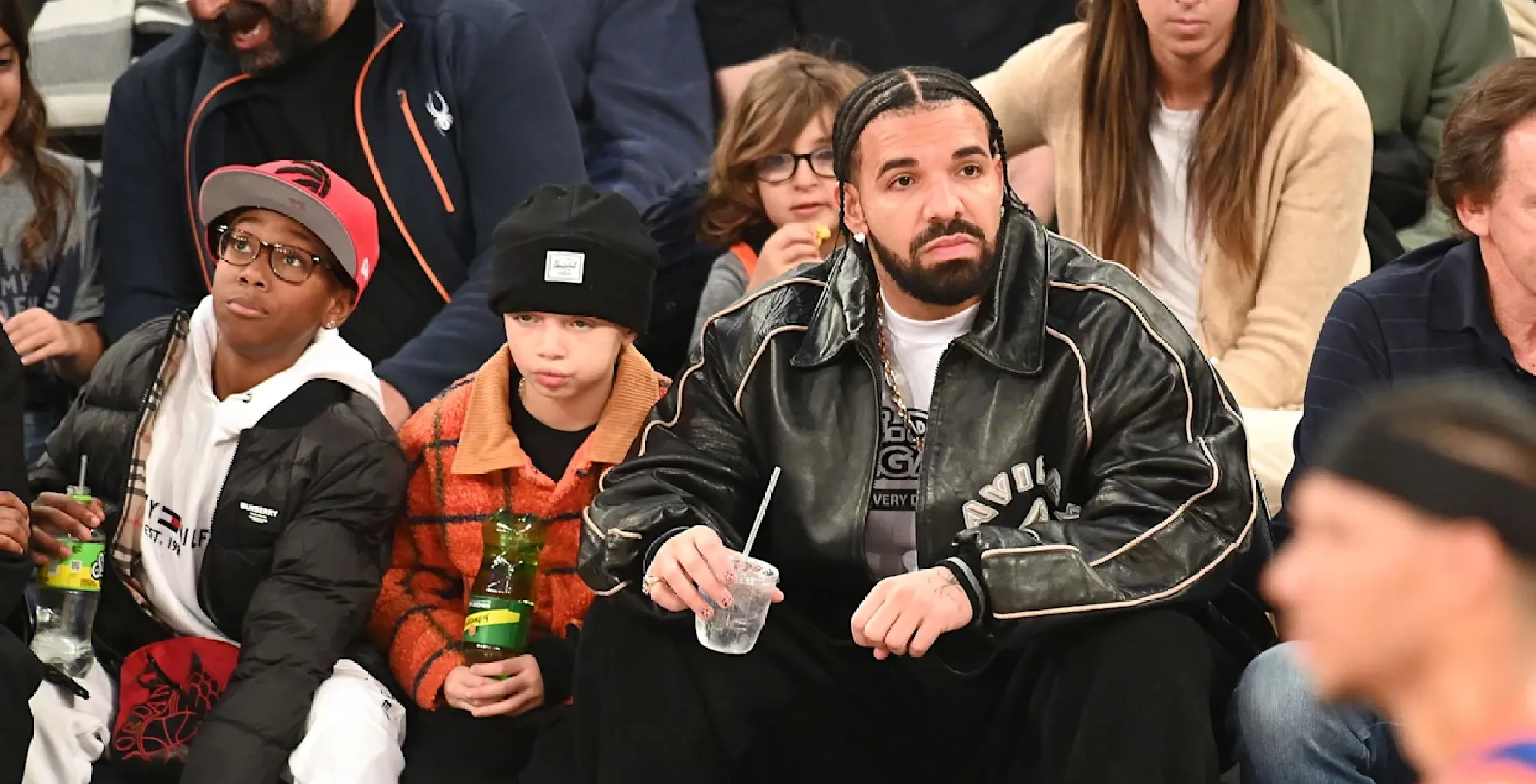 Drake wearing an oversized black distressed leather bomber jacket with Avirex 3D logo and beige piping at a Knicks game courtside