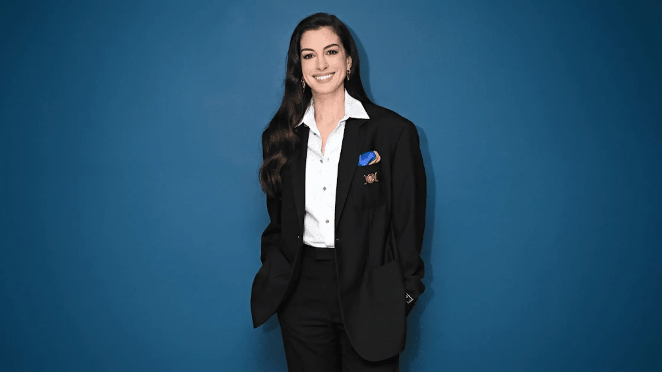 Anne Hathaway wearing an oversized black tailored Versace blazer with a blue and orange pocket square and a gold brooch on The Tonight Show Starring Jimmy Fallon.