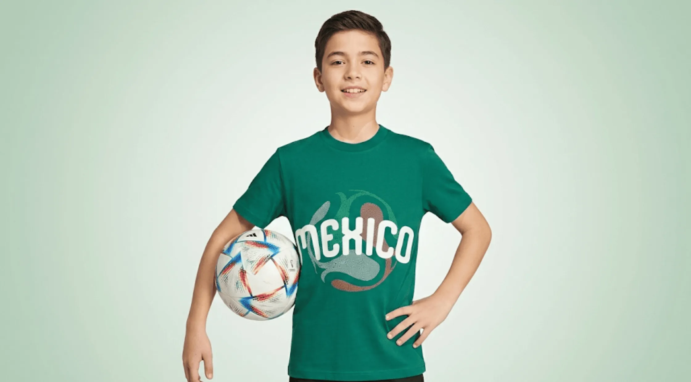Youth boy wearing the green FIFA World Cup 2026 Mexico graphic t-shirt while holding a soccer ball.