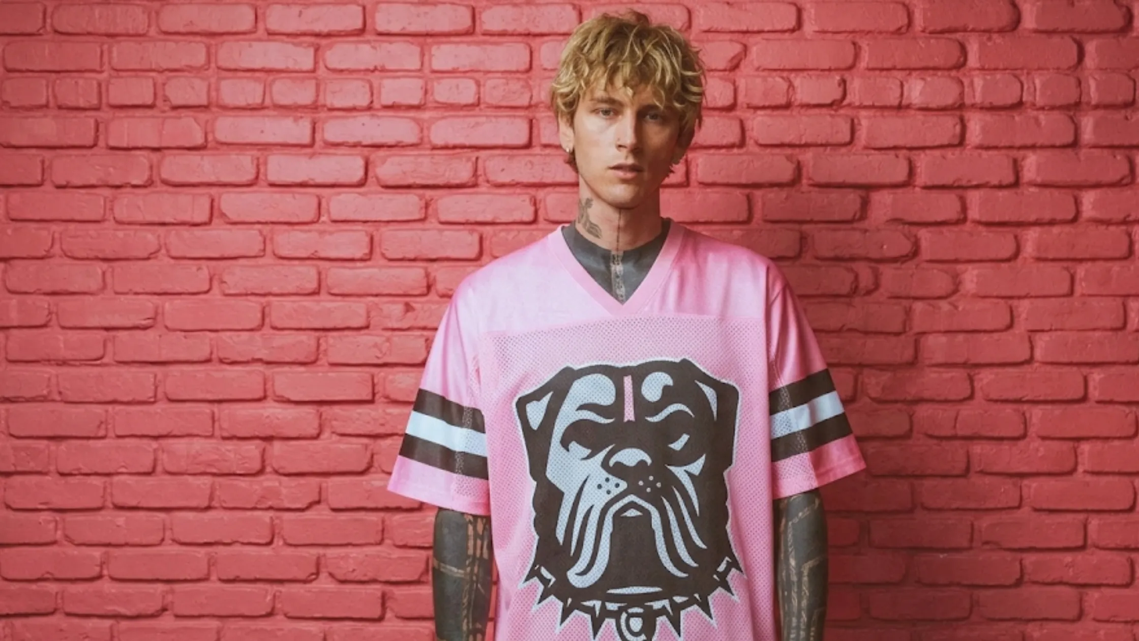 MGK X Cleveland Browns pink oversized football jersey featuring bulldog graphic, black and white varsity sleeve stripes, V-neck collar, and breathable mesh streetwear design.