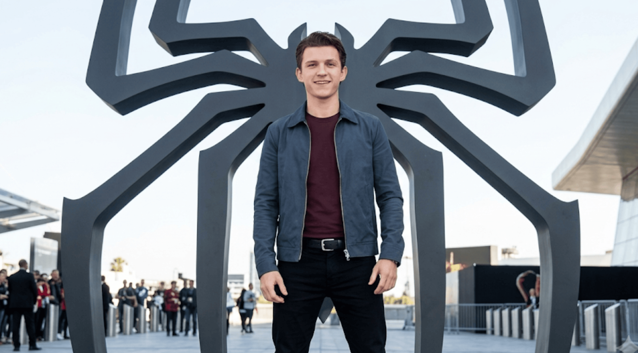 Tom Holland standing in front of a giant black Spider-Man logo wearing a slate blue Harrington zip jacket and a maroon shirt.