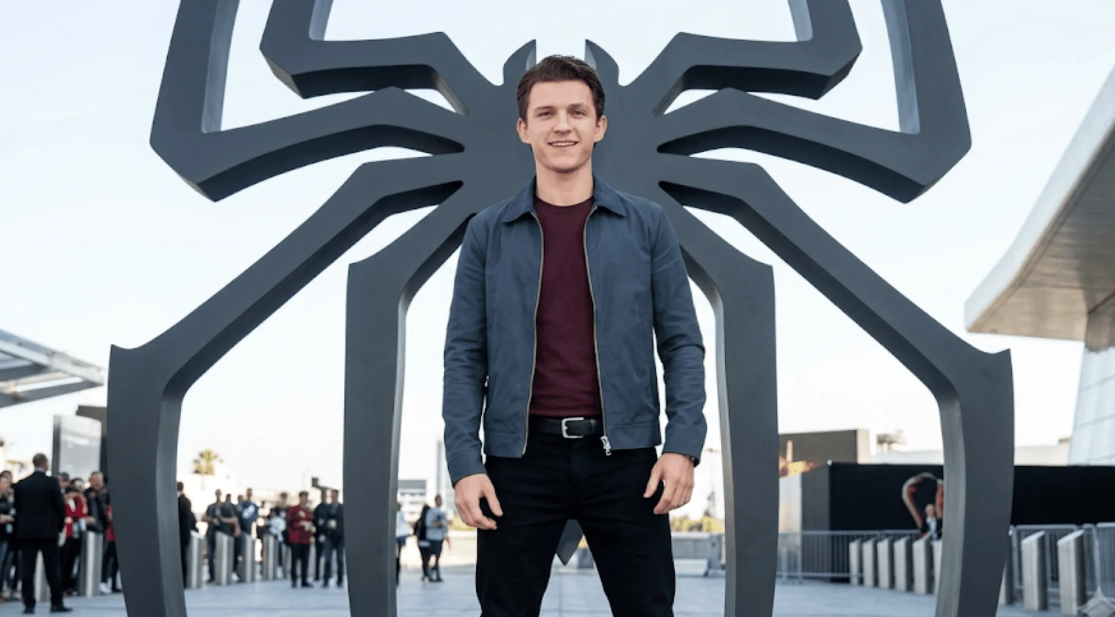 Tom Holland standing in front of a giant black Spider-Man logo wearing a slate blue Harrington zip jacket and a maroon shirt.