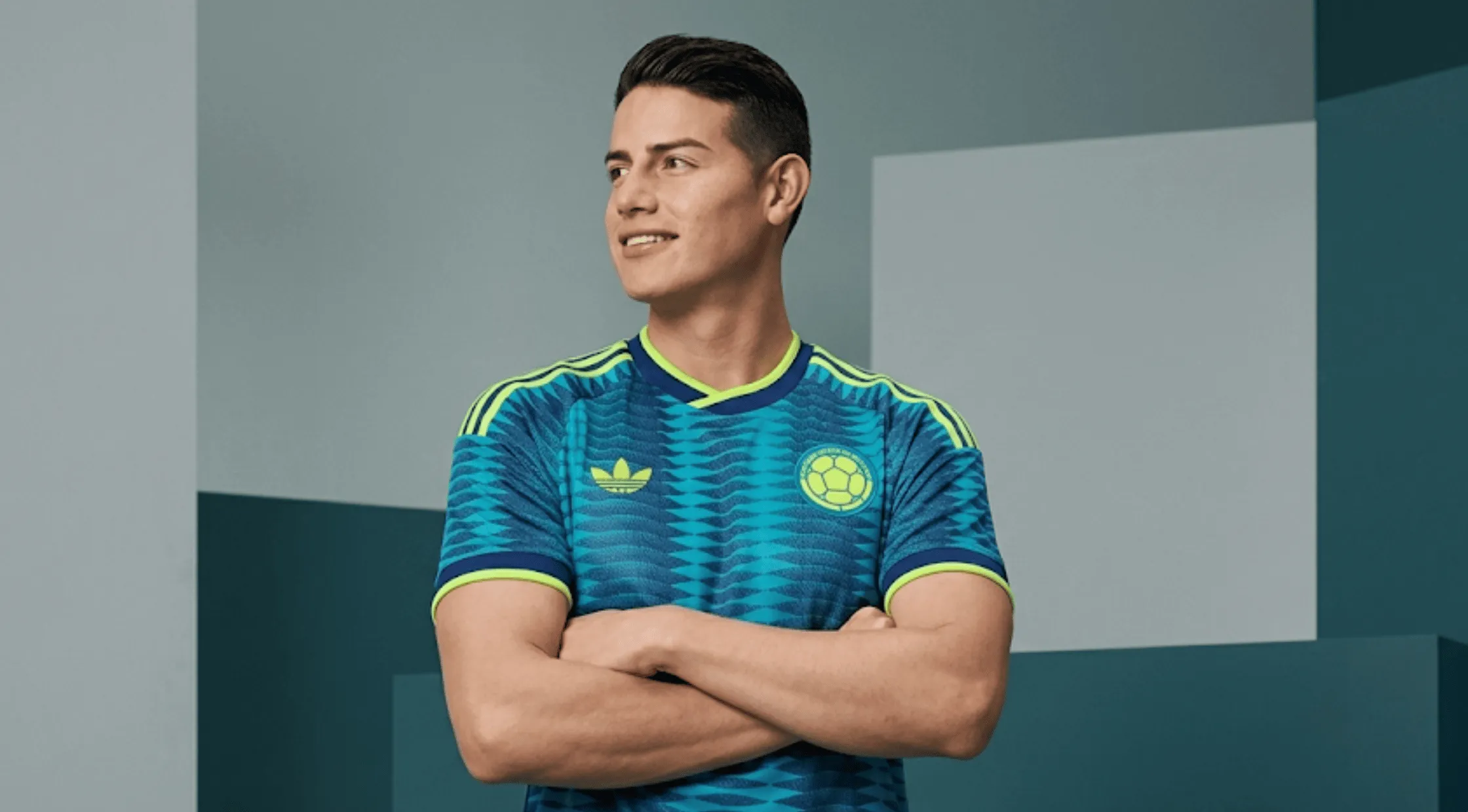 Colombian professional footballer James Rodríguez smiling and looking sideways with arms crossed, wearing the teal blue Colombia 2026 away jersey featuring a navy geometric wave pattern and neon lime green trim.
