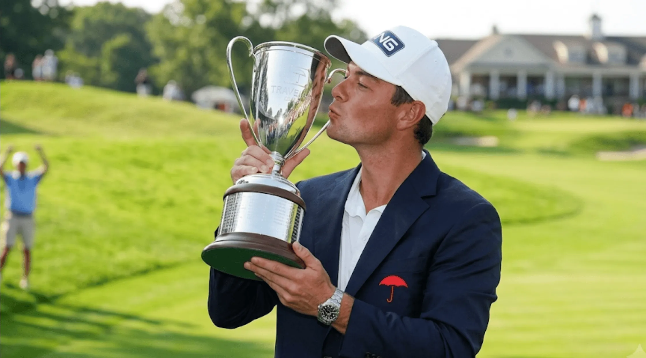 A close-up of professional golfer Viktor Hovland smiling on the green, wearing a navy blue single-breasted blazer adorned with an embroidered red umbrella logo on the left chest.