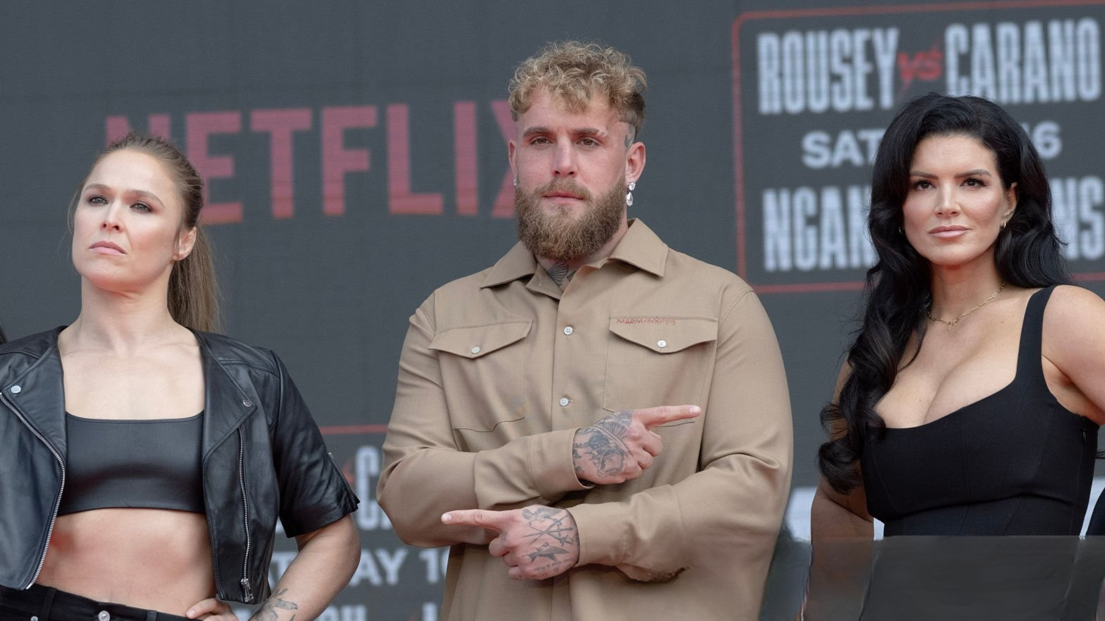 Ronda Rousey, Jake Paul, and Gina Carano facing off at the Intuit Dome press conference wearing a cropped leather jacket, tan overshirt outfit, and elegant black midi dress.