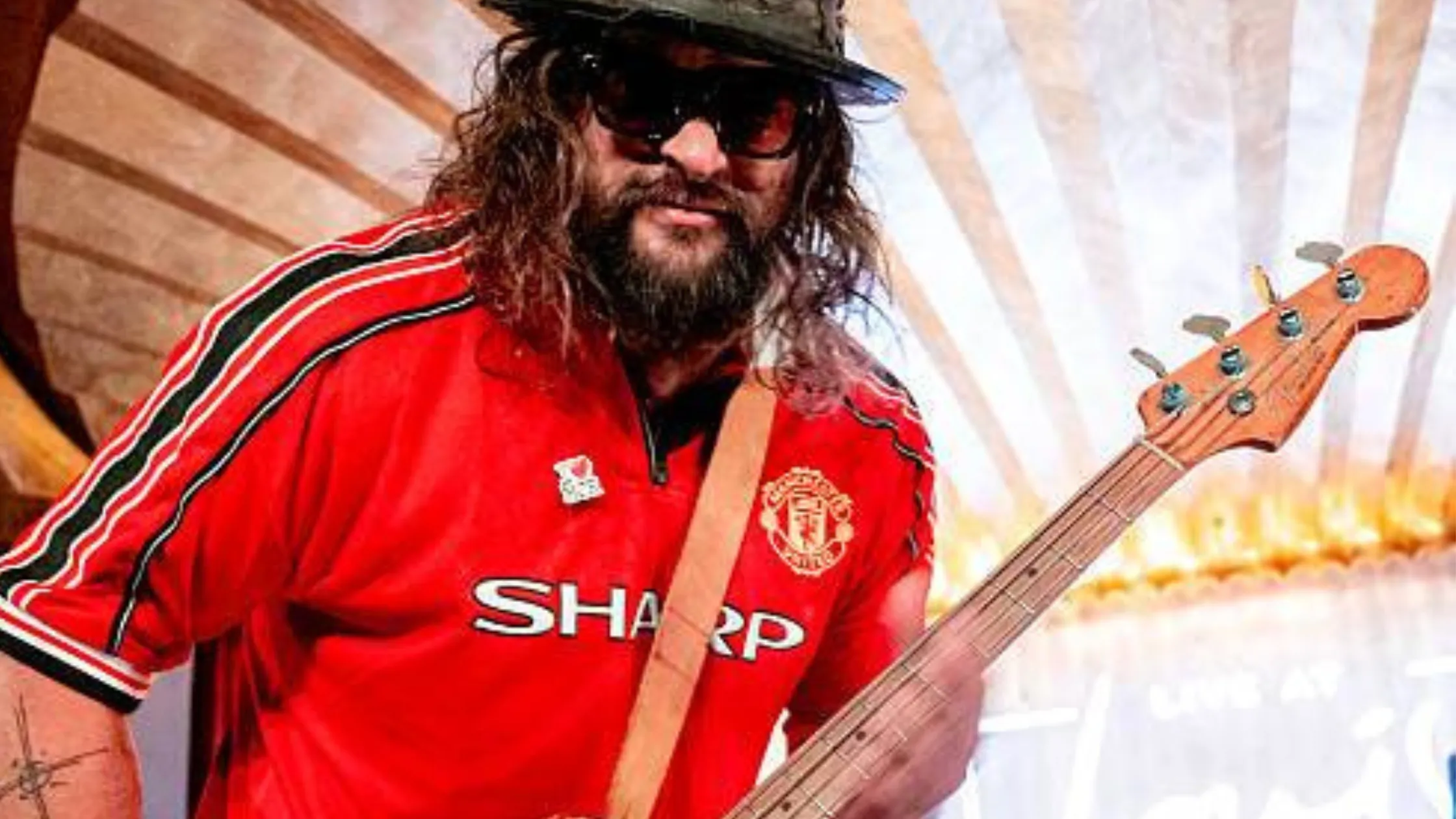 Oof Tatata stage star Jason Momoa wears a vibrant red Manchester United Oof Tatata T-shirt, adding retro football flair to his performance in Manchester.