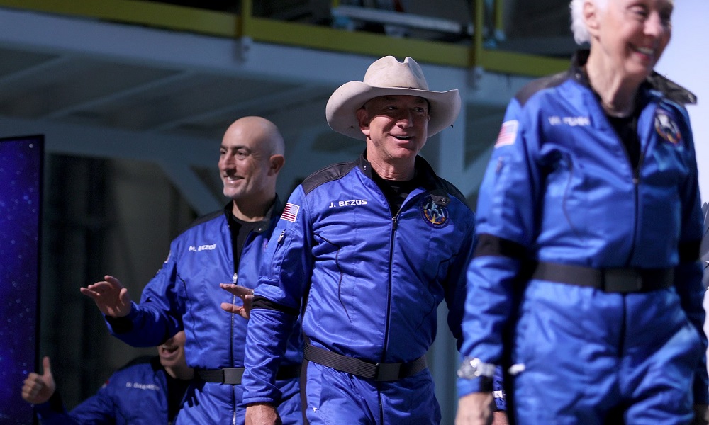 Jeff Bezos wearing royal blue Blue Origin space jumpsuit with black panels, mission patch, and US flag, striding in desert like a modern Chuck Yeager.