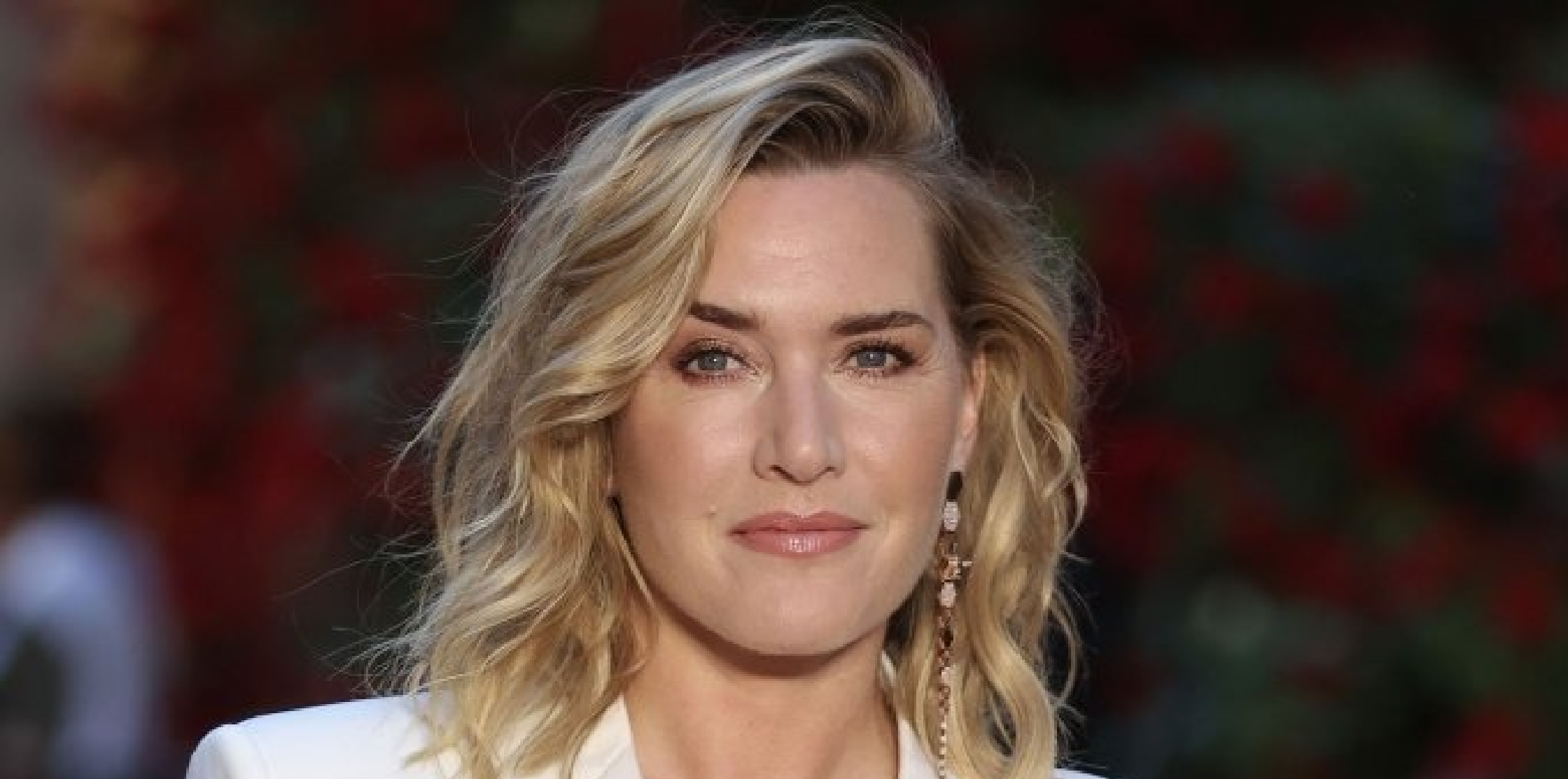 Kate Winslet walking in NYC wearing a mustard yellow single-breasted wool midi coat with patch pockets, holding a beige crossbody bag and a phone, styled with a cream top and natural makeup.