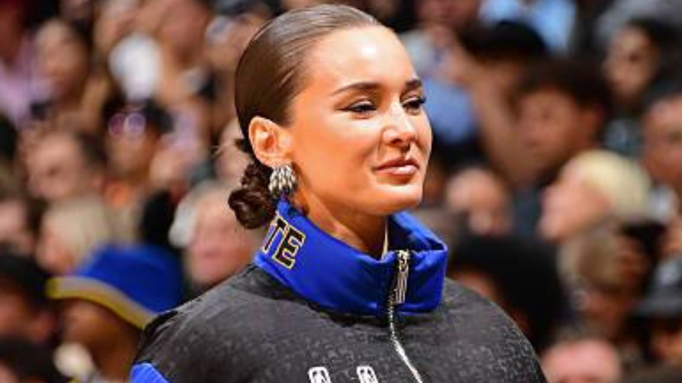 Kristin Juszczyk wears a bold blue, gold, and black Golden State Warriors puffer jacket with team logos and color-blocked design at Crypto.com Arena.