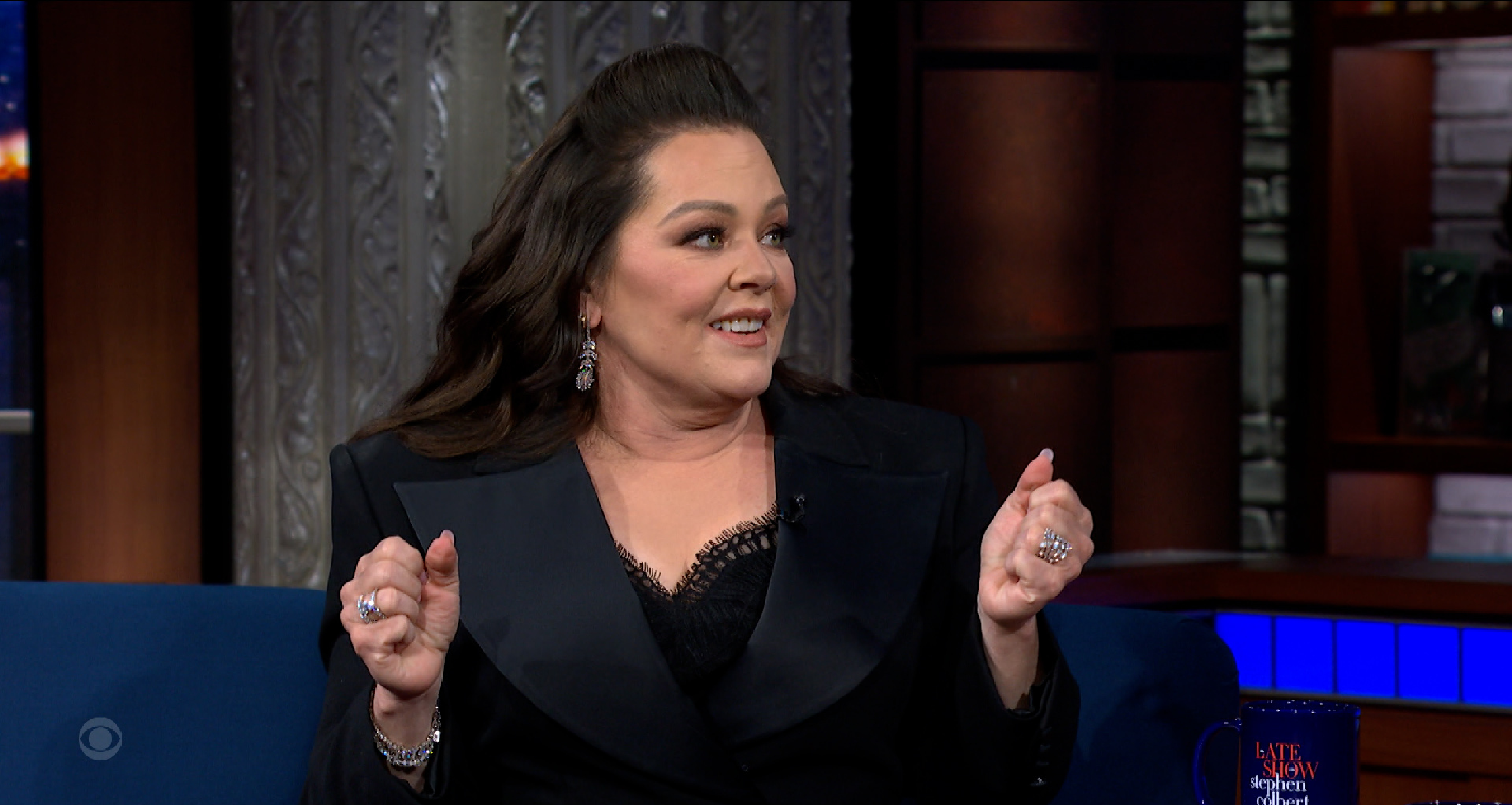 Melissa McCarthy walking in New York City outside The Late Show with Stephen Colbert wearing a long blue floral jacquard coat over a black dress, paired with embellished sheer ankle boots and carrying a black handbag.