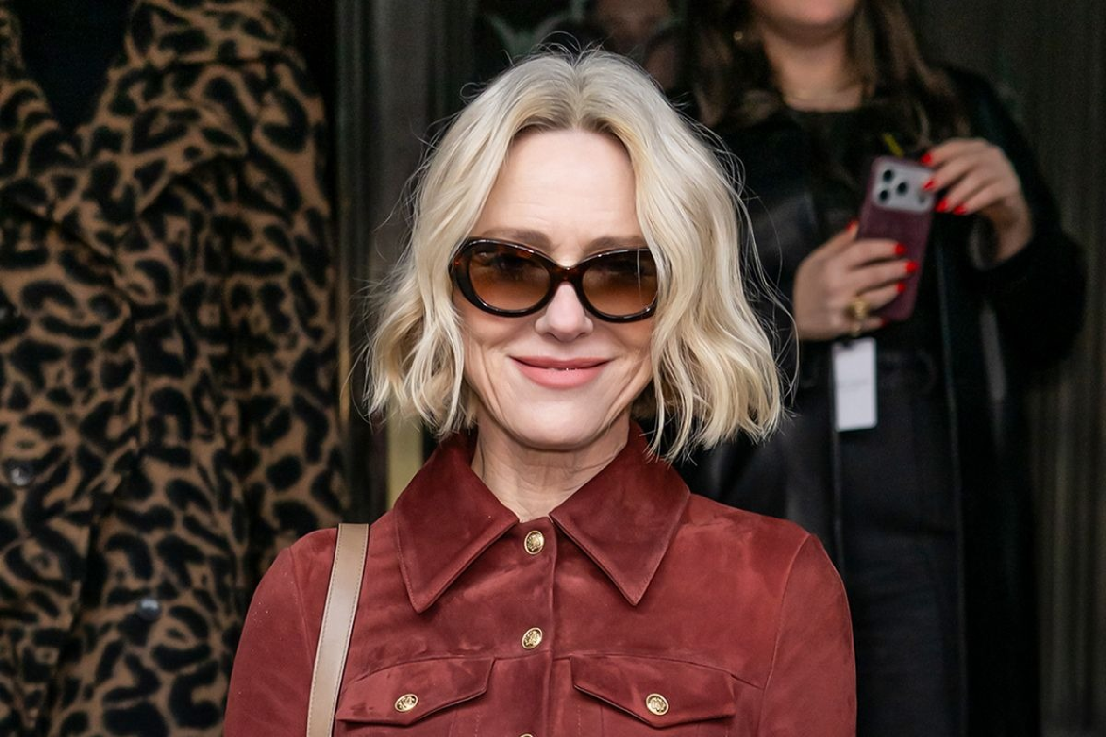Naomi Watts wearing a burgundy suede utility safari jacket with gold shank buttons and four flap pockets, paired with light blue wide-leg jeans at ALICE + OLIVIA Fall 2026 New York Fashion Week.