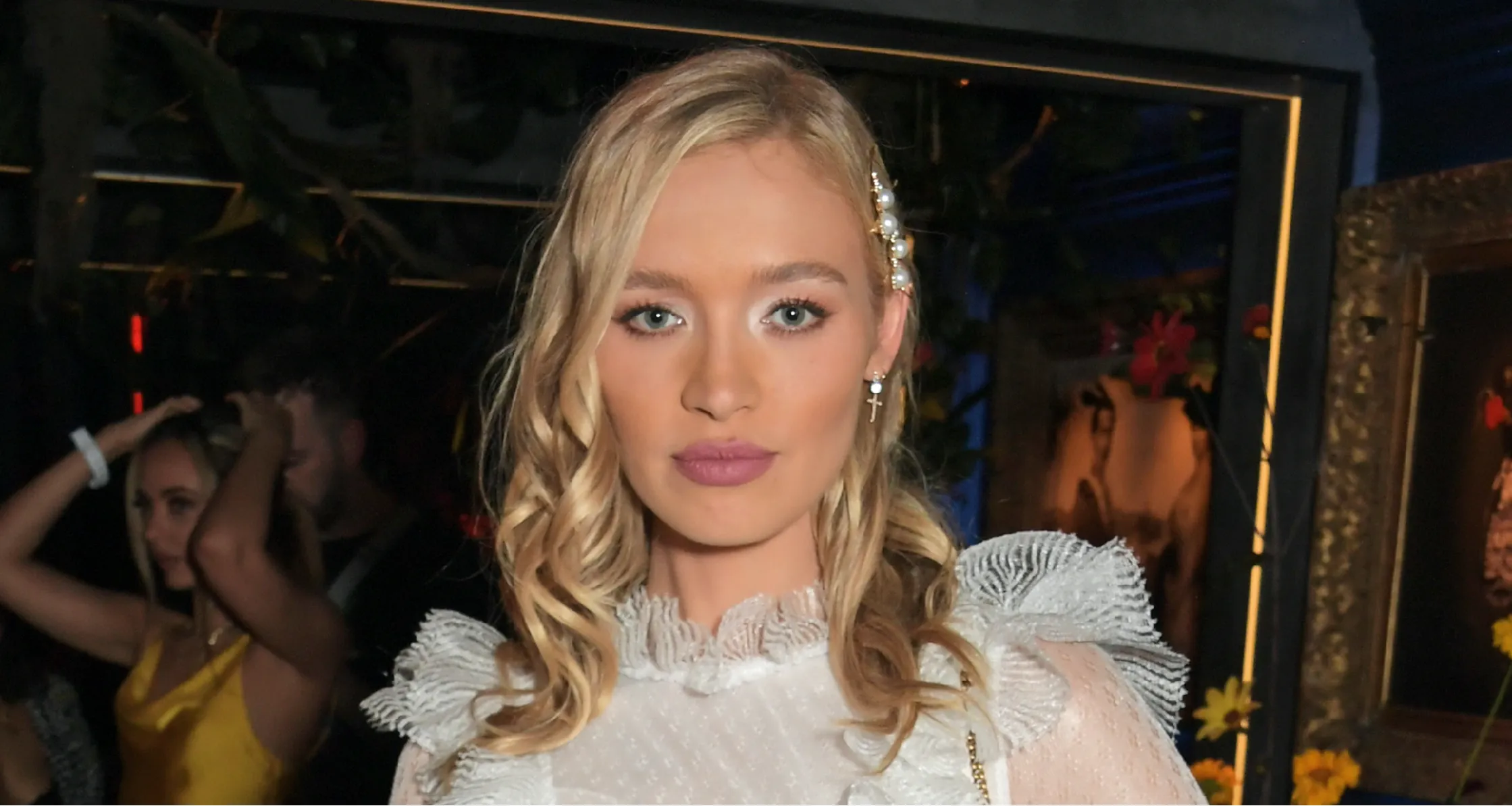 Roxy Horner wearing a taupe structured blazer with deep brown satin lapels over a white ruffled blouse, paired with black fitted trousers, styled with loose blonde waves and minimal evening makeup while arriving for a birthday night out in London.