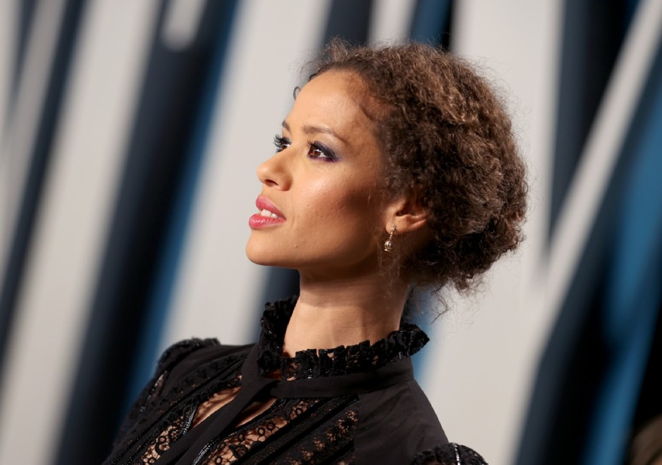 Gugu Mbatha-Raw wearing a long tailored black wool overcoat over a subtly shimmering black dress at the BAFTA x Vanity Fair party in London, paired with black heels, a matching clutch, and statement earrings, standing under warm evening event lighting.