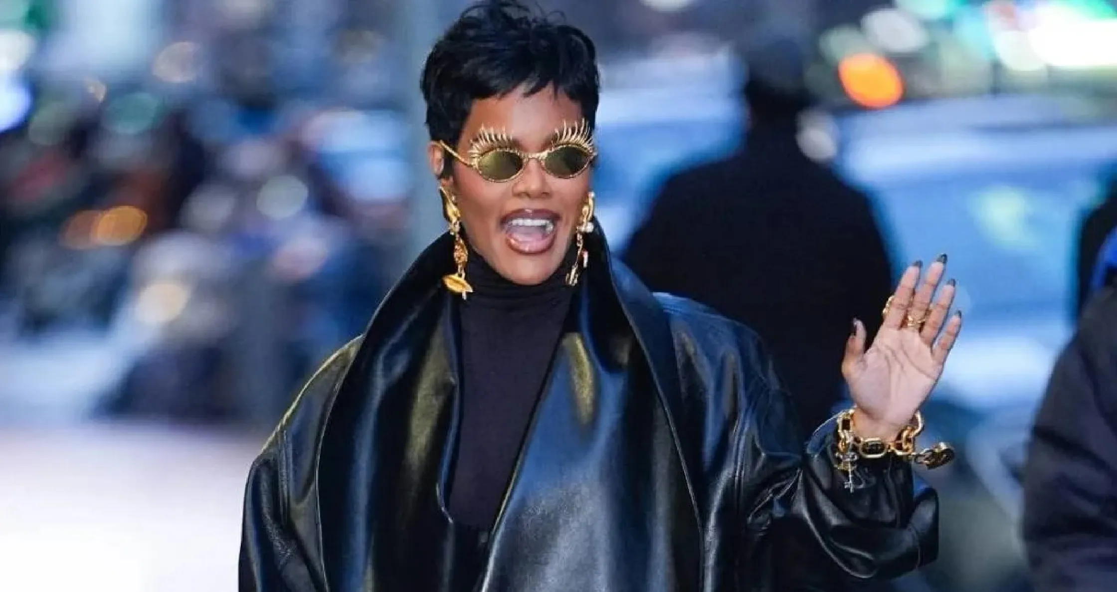 Teyana Taylor wearing the 2026 NYC black leather jacket with a dramatic oversized shawl collar, asymmetrical front wrap, balloon sleeves, and cinched waist, styled with high-waisted leather pants and gold “eye” sunglasses with spiked lashes.